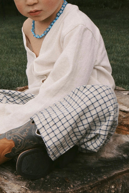 A child sits on a tree stump in a cream long-sleeve shirt and cabane childrenswear’s PER SE PANT blue check, paired with cowboy boots and a blue beaded necklace. Their face is partially visible, with grass in the background.