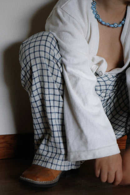 A person crouches indoors, showing only their lower face and torso, wearing cabane childrenswear’s PER SE PANT blue check with a white long-sleeve shirt, brown shoes, and a light blue beaded necklace.