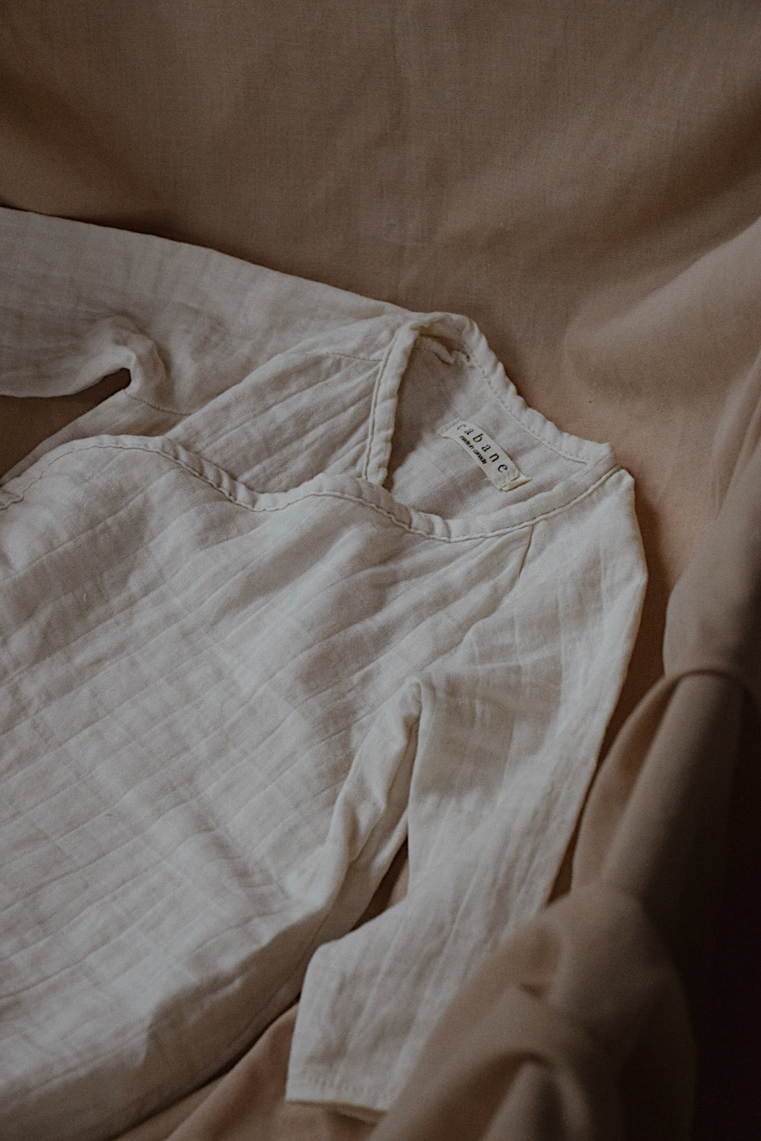 An Après Organic Wrap Undyed baby onesie from cabane childrenswear rests on a beige fabric backdrop. Crafted from breathable fabric, it features a small tag with text at the collar. The warm lighting enhances the cozy and serene essence, making it a perfect addition to any babies' layette collection.