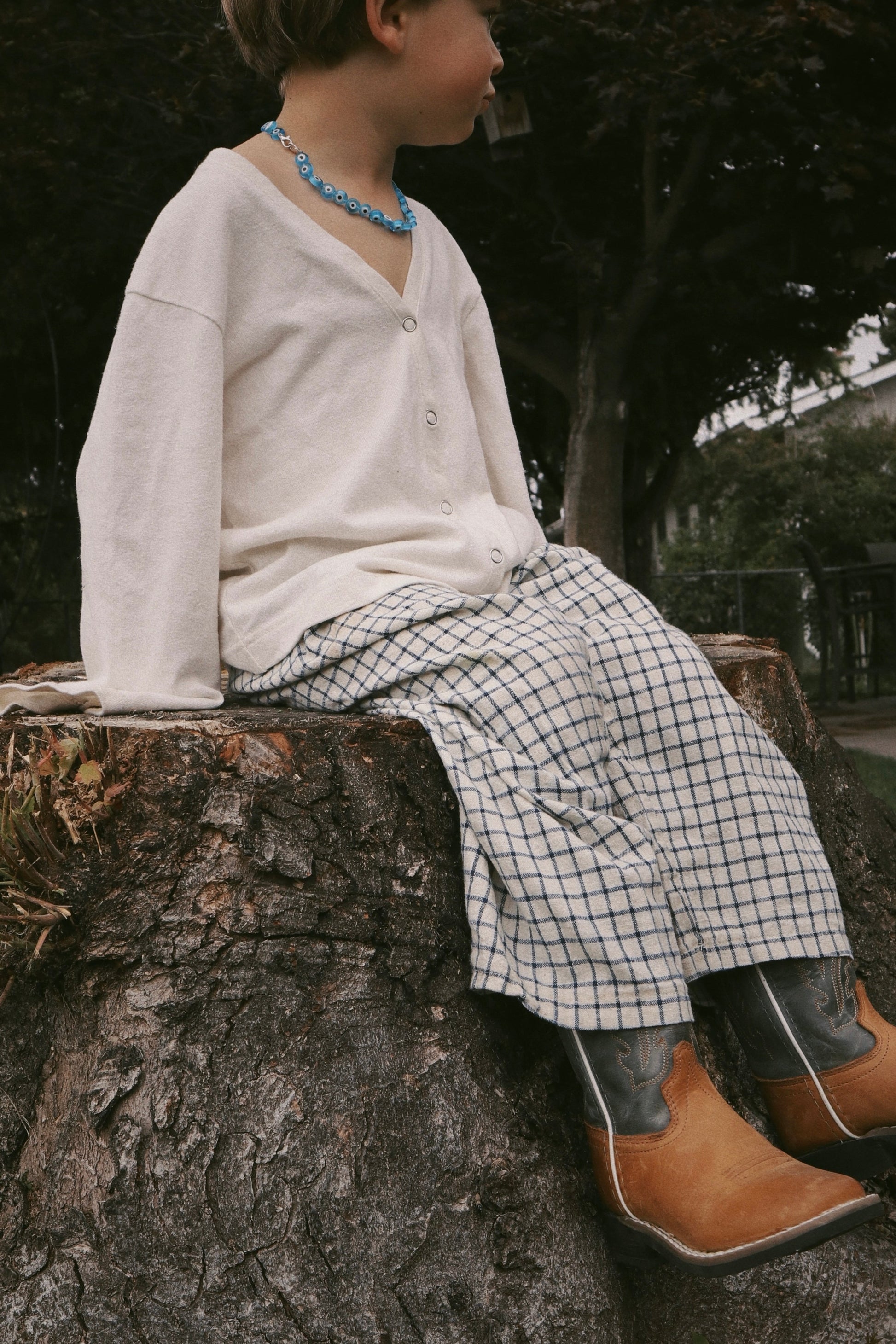 A child wearing cabane childrenswear's PER SE PANT blue check sits on a large tree stump outdoors, looking to the side.
