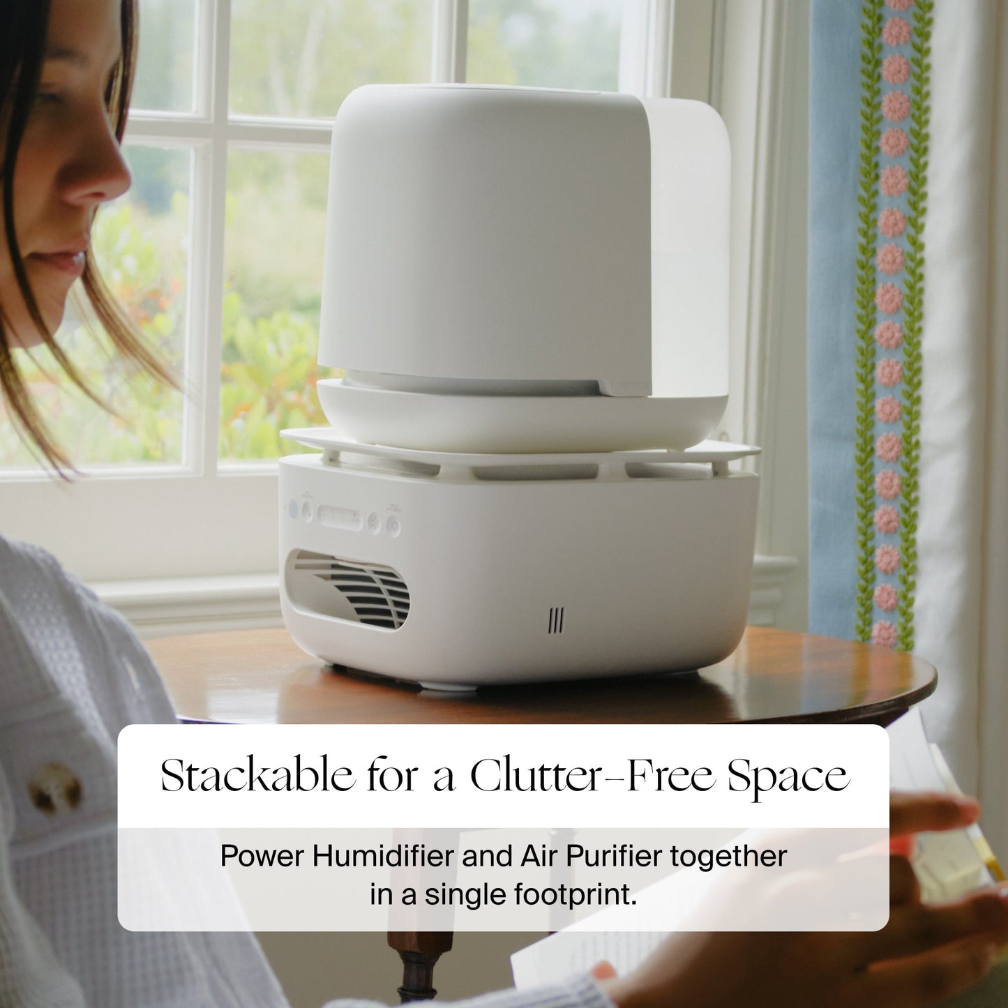 A woman sits by a window next to a white Canopy Nursery Air Purifier on the table—a device that combines humidifier and air purifier functions. Text overlay: "Stackable for a Clutter-Free Space. Humidify and purify in one with Canopy.
