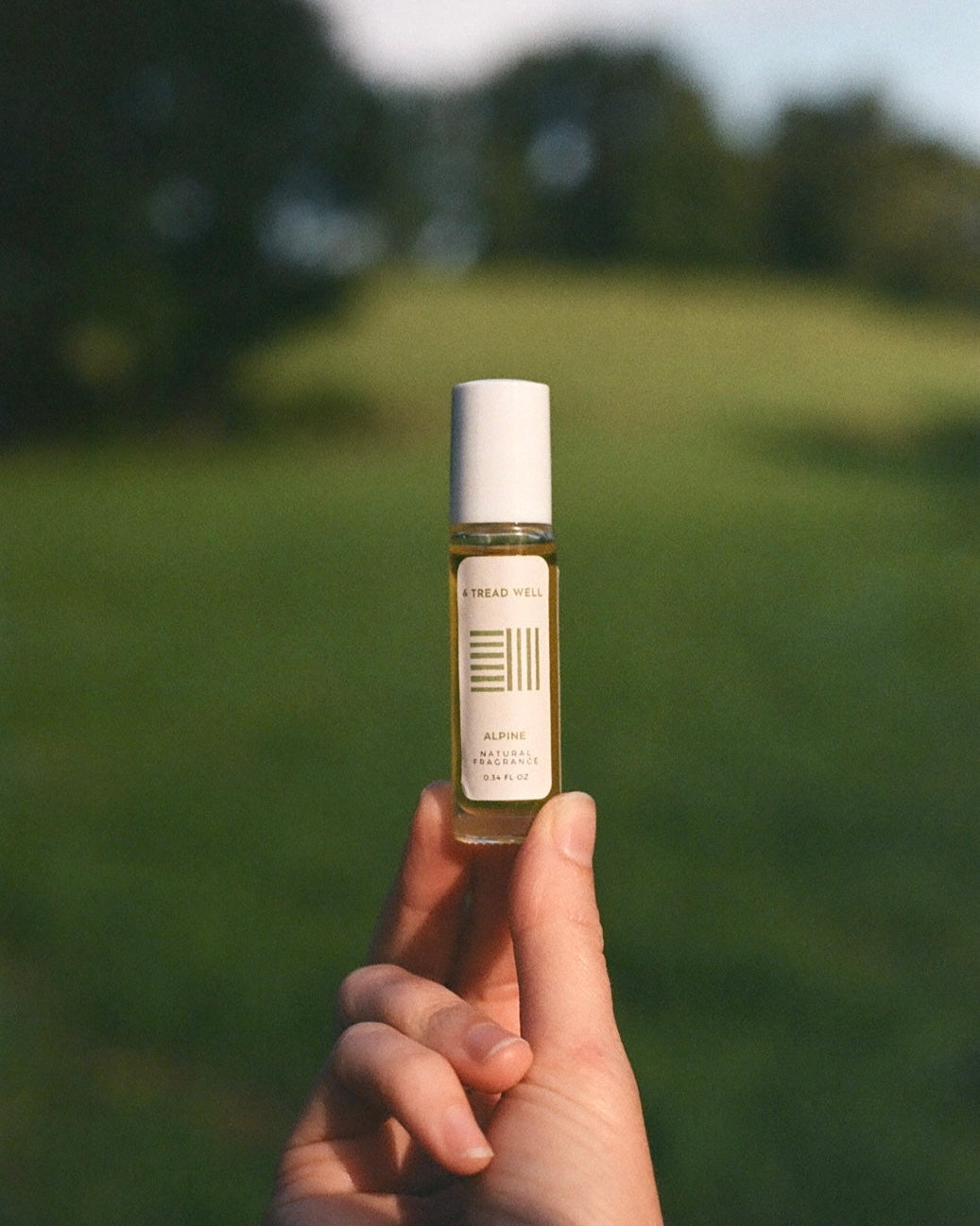 A hand holds a small glass bottle labeled "& Tread Well Natural Fragrance - Alpine" with a white cap. The unisex scent, made from natural ingredients, appears against softly blurred green grass and trees in natural light.