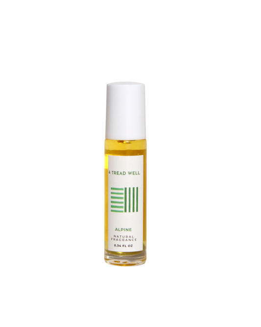 A small glass bottle with a white cap and roll-on applicator contains yellow "Natural Fragrance - Alpine" by & Tread Well (0.34 FL OZ), featuring organic essential oils and a label with green design elements.