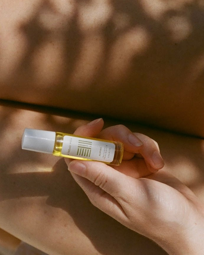 A hand holds a small glass bottle of & Tread Well’s Natural Fragrance - Alpine, its white label and cap glowing in gentle sunlight on bare skin, showcasing this unisex scent crafted from natural ingredients for a pure aesthetic.