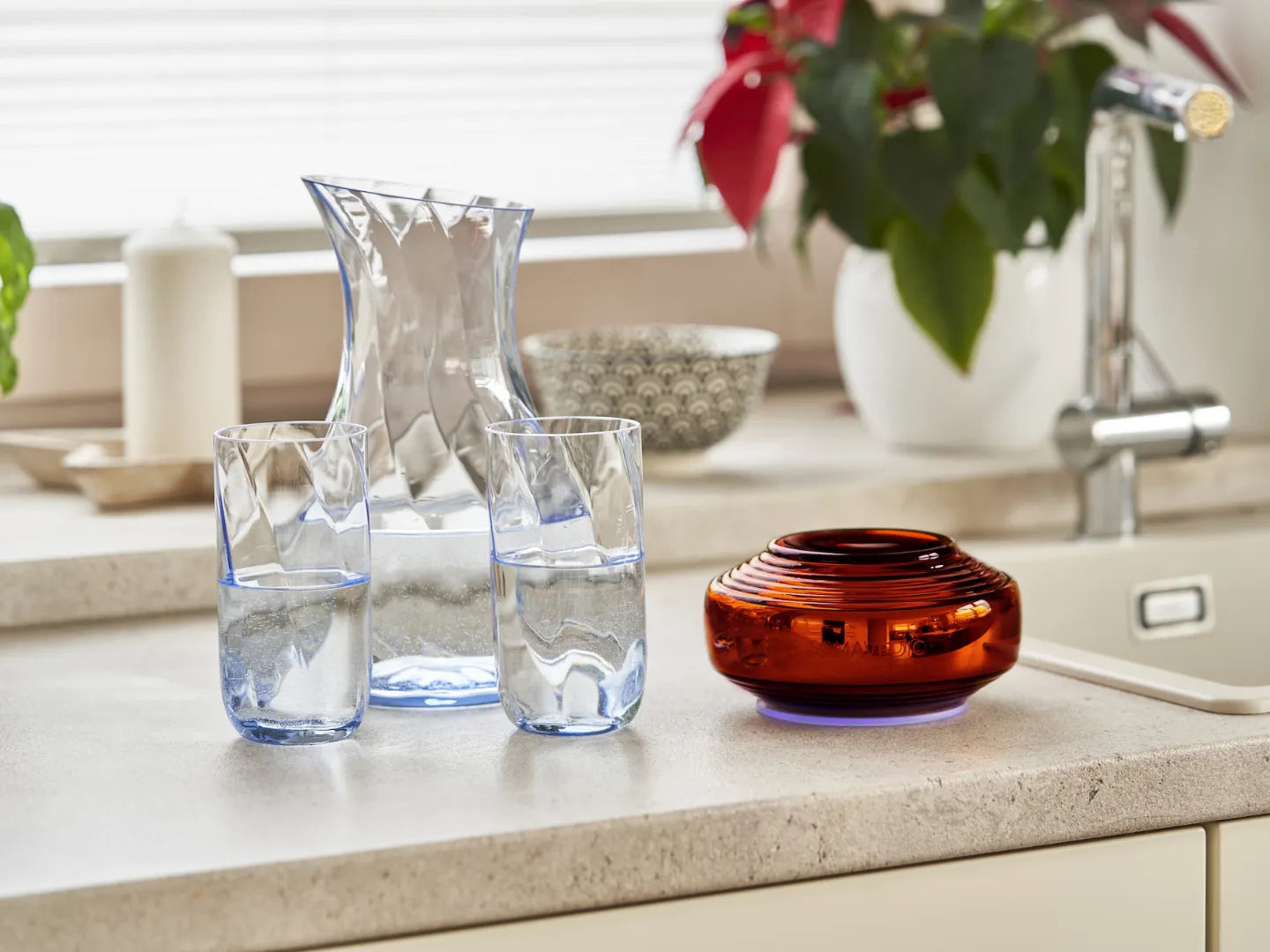 A clear jug and two filled glasses are placed on a kitchen countertop next to Somavedic USA's glossy Amber vase. In the background, plants, a patterned bowl, and a faucet enhance the cozy atmosphere—creating an ideal space for ultimate EMF protection.