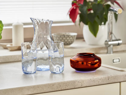 A clear jug and two filled glasses are placed on a kitchen countertop next to Somavedic USA's glossy Amber vase. In the background, plants, a patterned bowl, and a faucet enhance the cozy atmosphere—creating an ideal space for ultimate EMF protection.