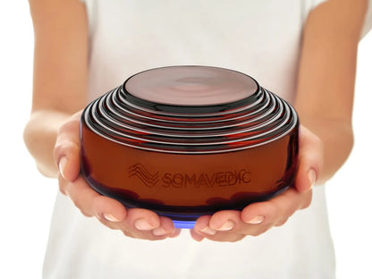 A person is holding a smooth, dome-shaped, orange-brown device labeled "Somavedic USA Amber" against a plain white background, designed to provide ultimate EMF protection and mitigate 5G effects.