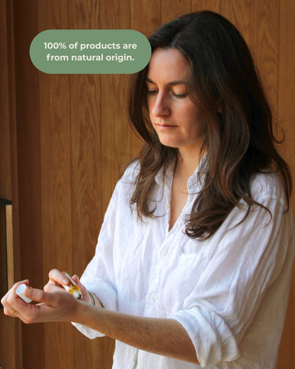 A woman with long brown hair in a white shirt applies & Tread Well's Natural Fragrance - Nectar to her hand. A green bubble reads, "100% of products are from natural origin." She stands before wood, surrounded by sweet orange and cardamom scent.