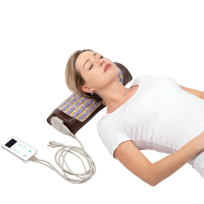 A woman is lying down with her head resting on a HealthyLine Amethyst Bolster Firm | Heated InfraMat Pro®. The device, featuring a patterned brown surface and a control panel attached via a cord, promotes natural healing. She appears relaxed with eyes closed, wearing a white shirt and a delicate necklace.