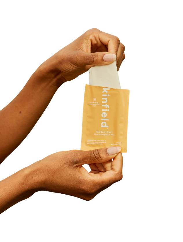 Two hands open a yellow Kinfield Golden Hour™ Wipes packet, pulling out a mosquito repellent wipe infused with Indonesian citronella, set against a plain background.