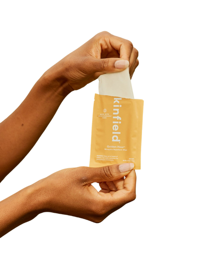 Two hands open a yellow Kinfield Golden Hour™ Wipes packet, pulling out a mosquito repellent wipe infused with Indonesian citronella, set against a plain background.