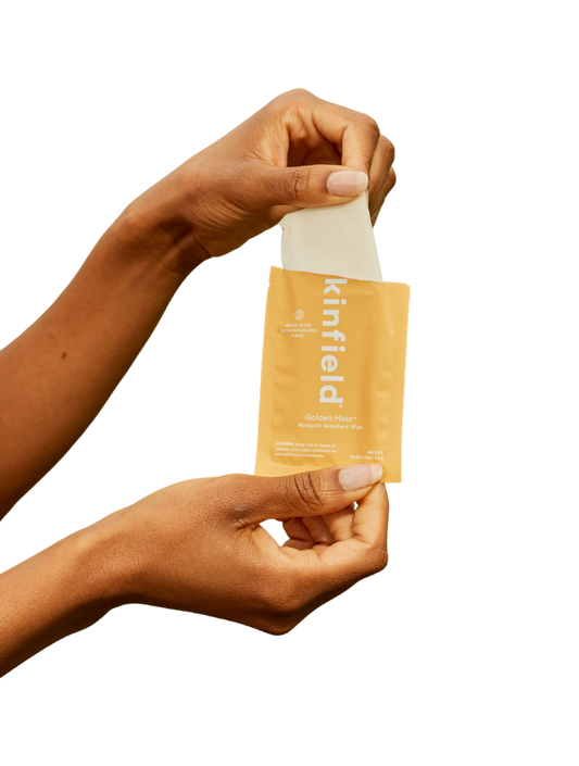 Two hands open a yellow Kinfield Golden Hour™ Wipes packet, pulling out a mosquito repellent wipe infused with Indonesian citronella, set against a plain background.