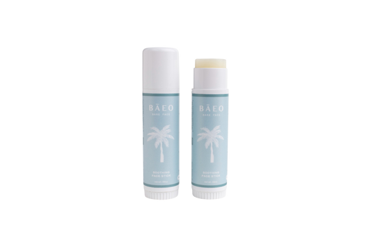 Two light blue tubes of bāeo bare face hydrating stick, with white palm tree graphics—one open to reveal the balm, one closed—are shown on a white background. Made with organic botanicals for gentle skin care.