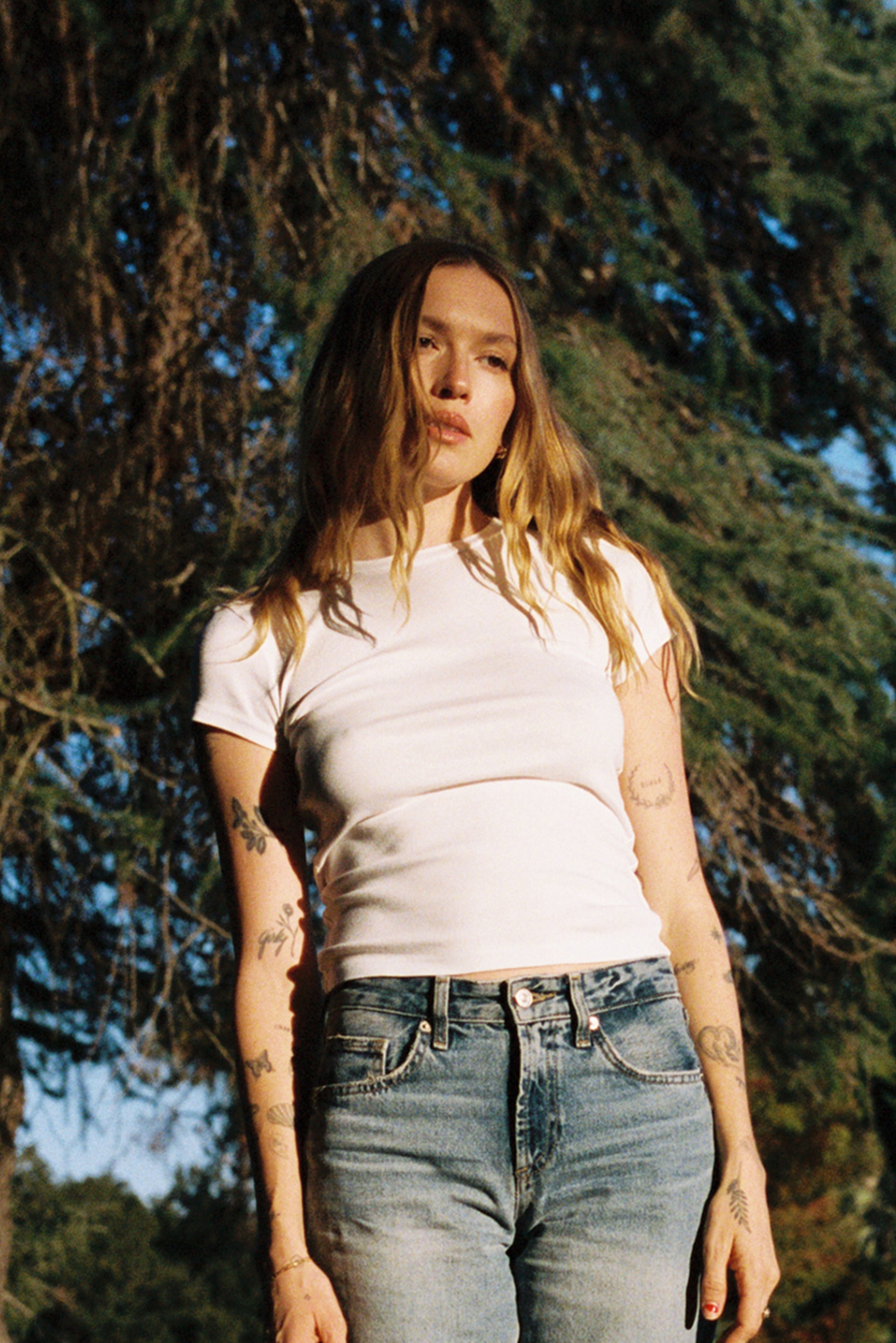 White A person with long hair stands outdoors in front of a tree, wearing the LA Relaxed Bebe Organic Tee and jeans. Tattoos are visible on their arms. Sunlight casts a warm glow, enhancing the natural surroundings and complementing the tee's organic cotton feel.
