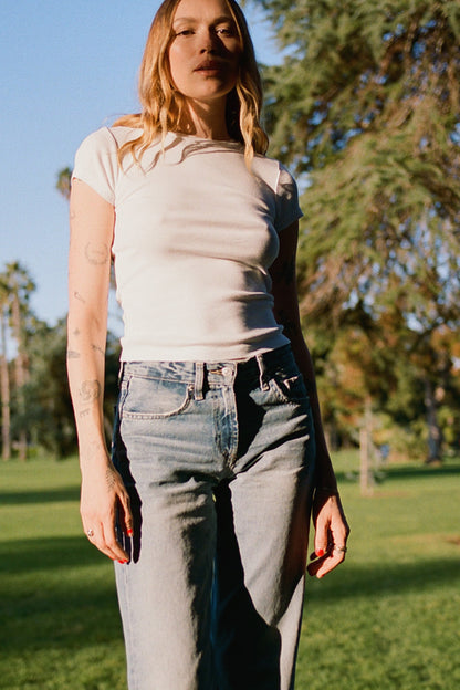 White A person with long hair, wearing a Bebe Organic Tee by LA Relaxed and jeans, stands in a sunlit park. Their tattooed arms are highlighted against a backdrop of trees and vibrant green grass, creating a serene scene.