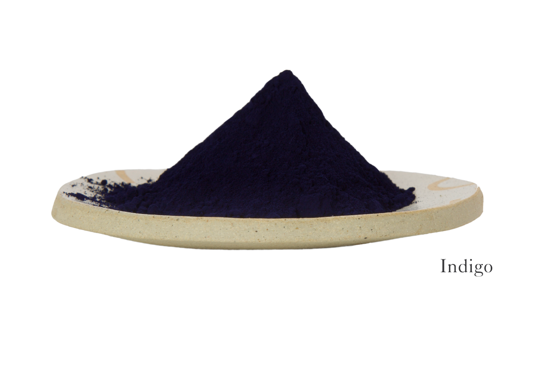 A mound of dark blue powder labeled "Indigo" sits on a beige ceramic dish against a white background, evocative of the deep hue featured in the Indigo Ayurvedic Yoga Mat by Bennd Yoga.