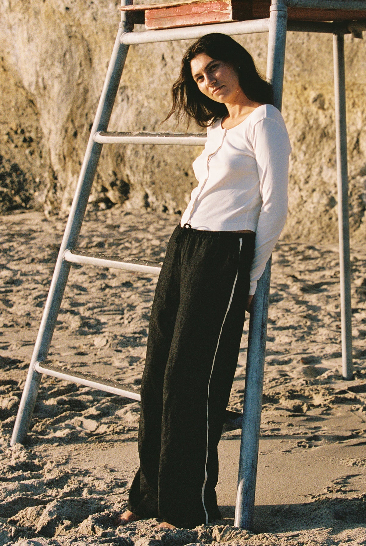A woman in a white long-sleeve top and LA RELAXED Linen Track Pant with a drawstring waistband leans casually against a metal lifeguard ladder on a sandy beach, with rocky cliffs in the background.