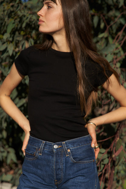 Black A person with long brown hair is outdoors, wearing an LA Relaxed Bebe Organic Tee and blue jeans. They gaze to the side with hands on hips, surrounded by greenery.