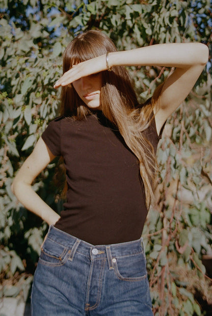 Black A woman with long hair, wearing a Bebe Organic Tee by LA Relaxed and jeans, stands outdoors with her hand shielding her eyes from the sunlight. She poses in front of leafy greenery, creating a casual, sunlit scene.