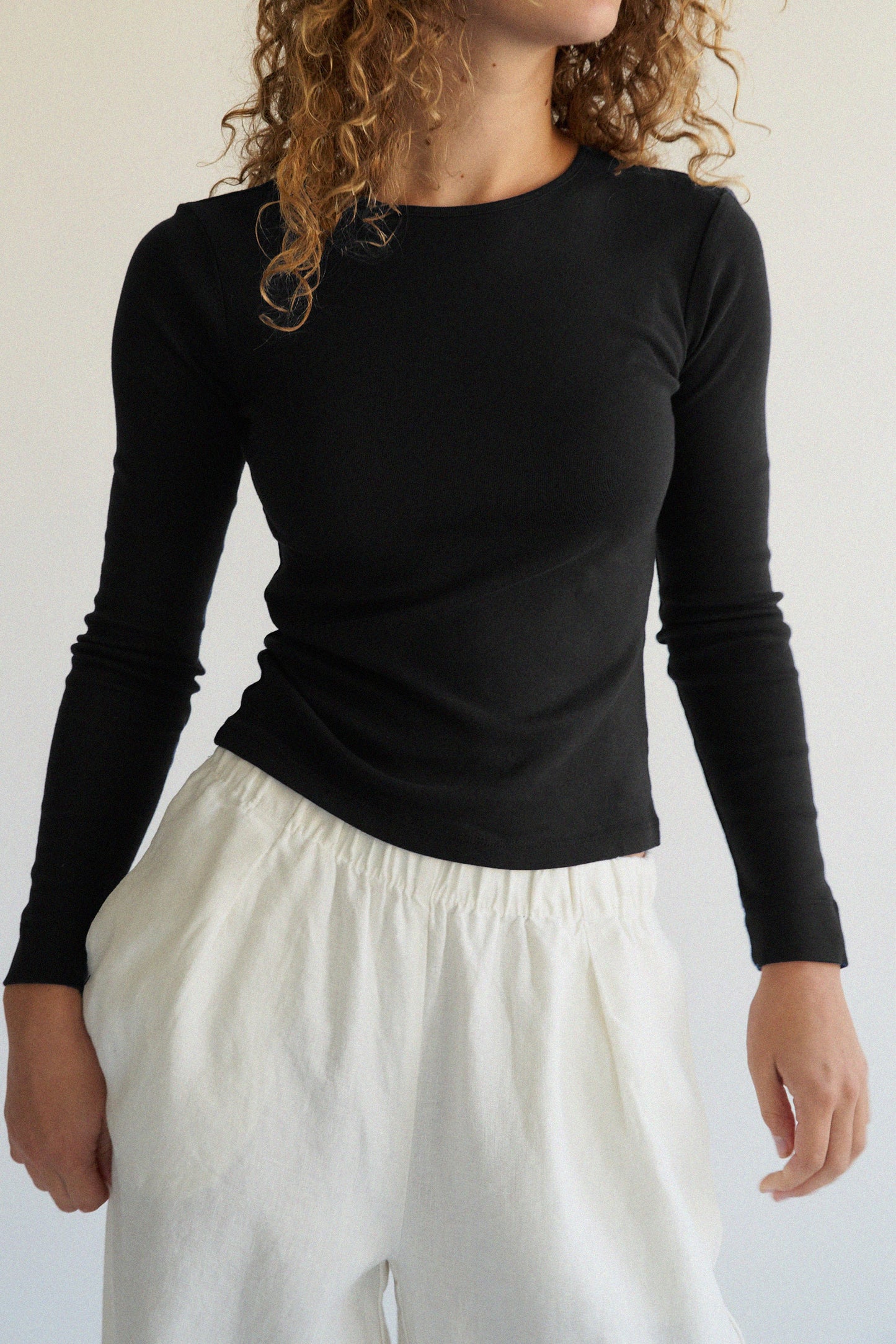 A person with curly hair elegantly stands against a neutral background wearing an LA RELAXED Bebe Organic Long Sleeve in black and white pants.