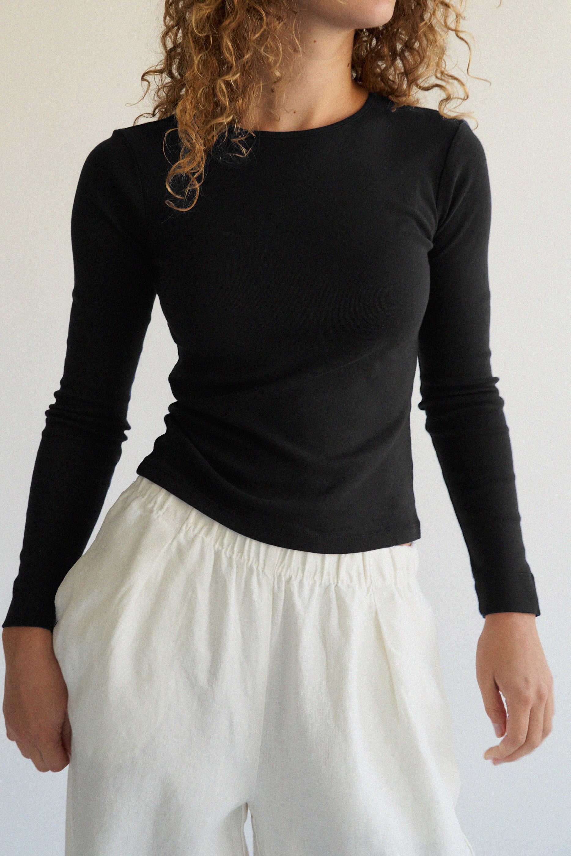 A person with curly hair elegantly stands against a neutral background wearing an LA RELAXED Bebe Organic Long Sleeve in black and white pants.