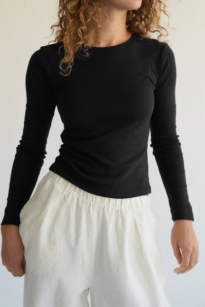 A person with curly hair elegantly stands against a neutral background wearing an LA RELAXED Bebe Organic Long Sleeve in black and white pants.