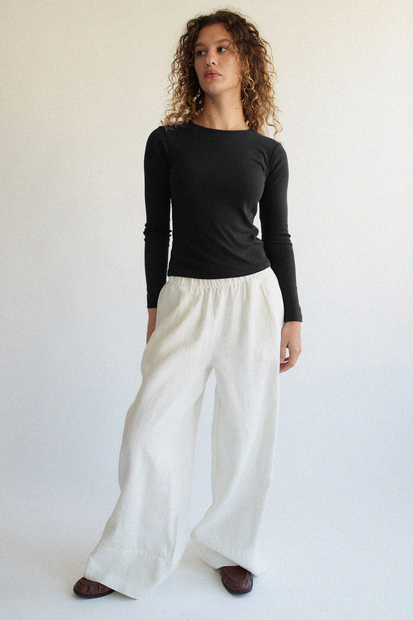 A person with curly hair, wearing a Bebe Organic Long Sleeve by LA RELAXED and white wide-legged pants, stands against a plain light background. They look slightly to the side with a neutral expression.
