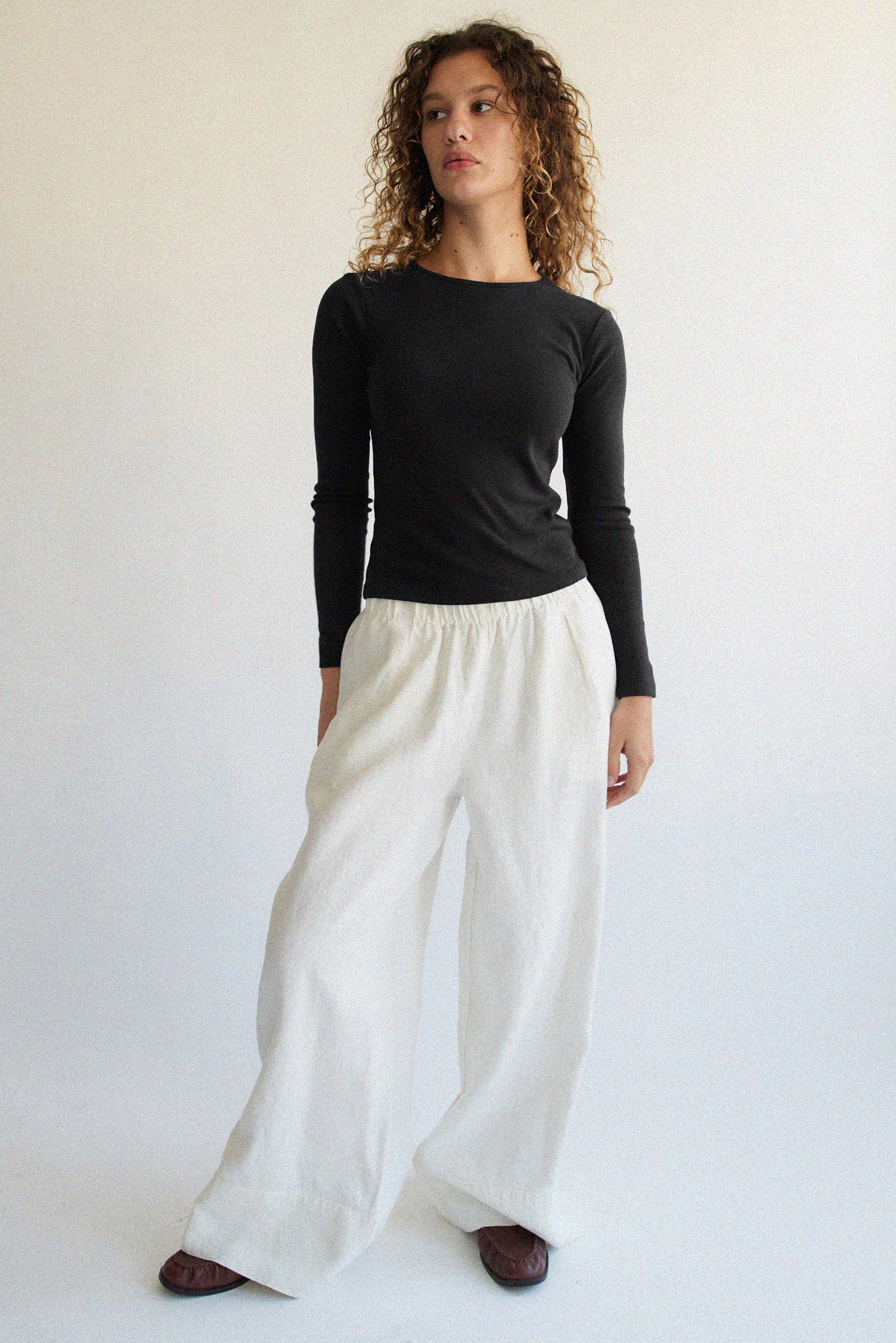 A person with curly hair, wearing a Bebe Organic Long Sleeve by LA RELAXED and white wide-legged pants, stands against a plain light background. They look slightly to the side with a neutral expression.
