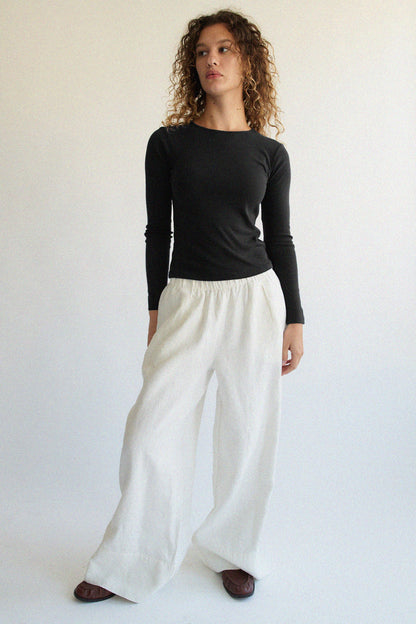 A person with curly hair, wearing a Bebe Organic Long Sleeve by LA RELAXED and white wide-legged pants, stands against a plain light background. They look slightly to the side with a neutral expression.