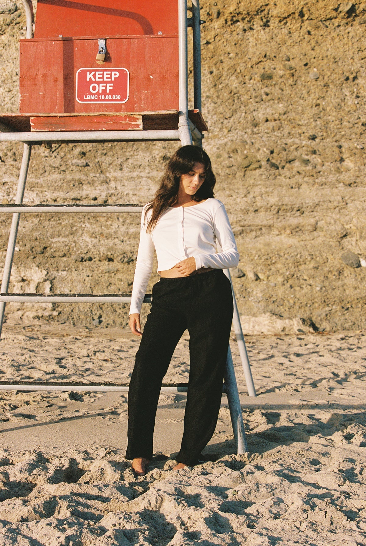 A woman wears LA RELAXED’s Linen Track Pant—black with a drawstring waistband—on a sandy beach in front of a red “KEEP OFF” lifeguard stand, rocky cliffs rising in the background.