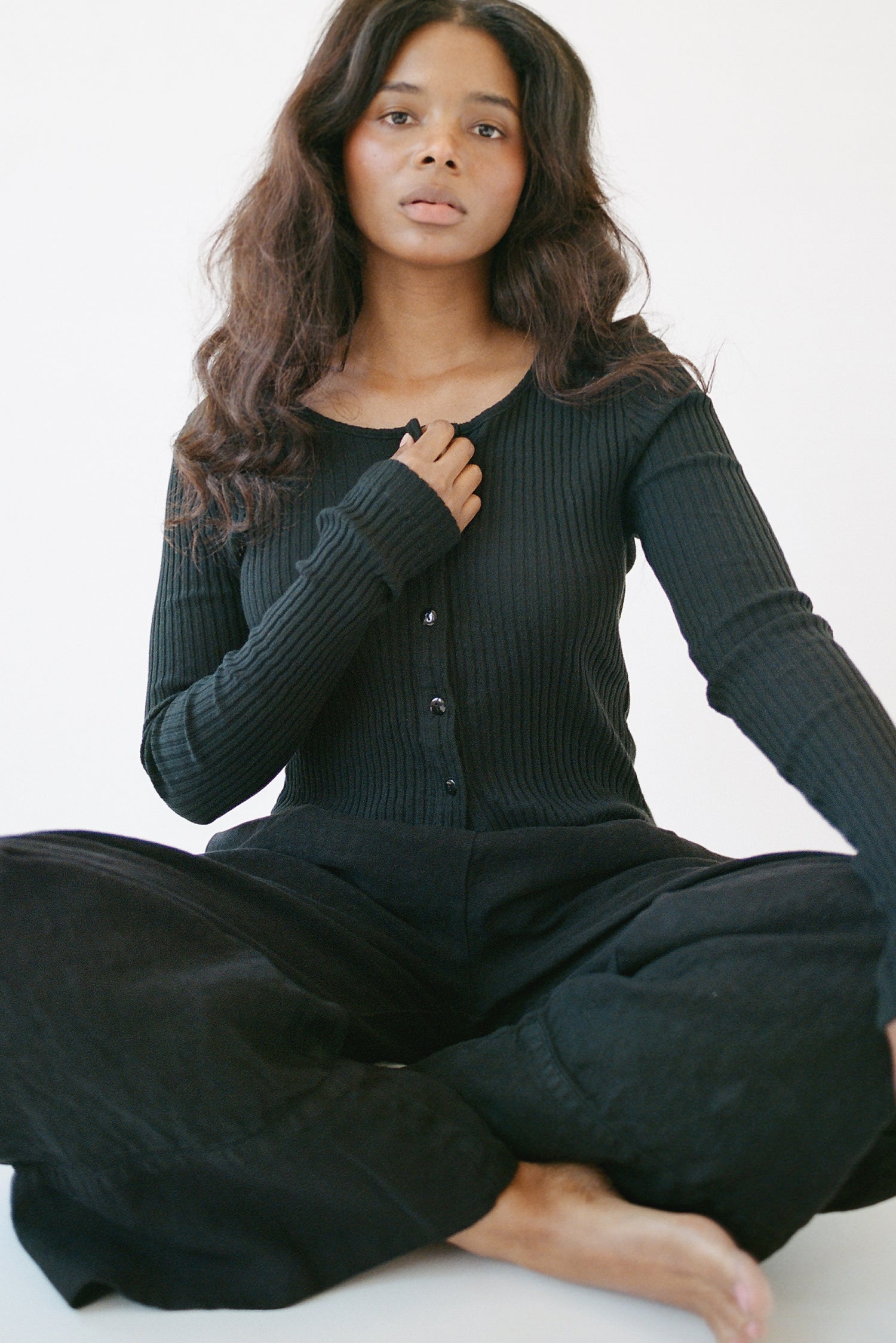 Black - 
A woman with long wavy hair sits cross-legged on the floor against a white background, wearing the LA RELAXED Pointelle Cardigan with pearlescent buttons and loose black pants, one hand on her chest and a calm expression.