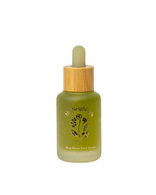 A green glass dropper bottle with a wooden cap, labeled "My Neighbor’s Tallow Blue Moon Face Serum" and adorned with a floral design, contains this organic serum for sensitive skin. The product is shown on a plain white background.
