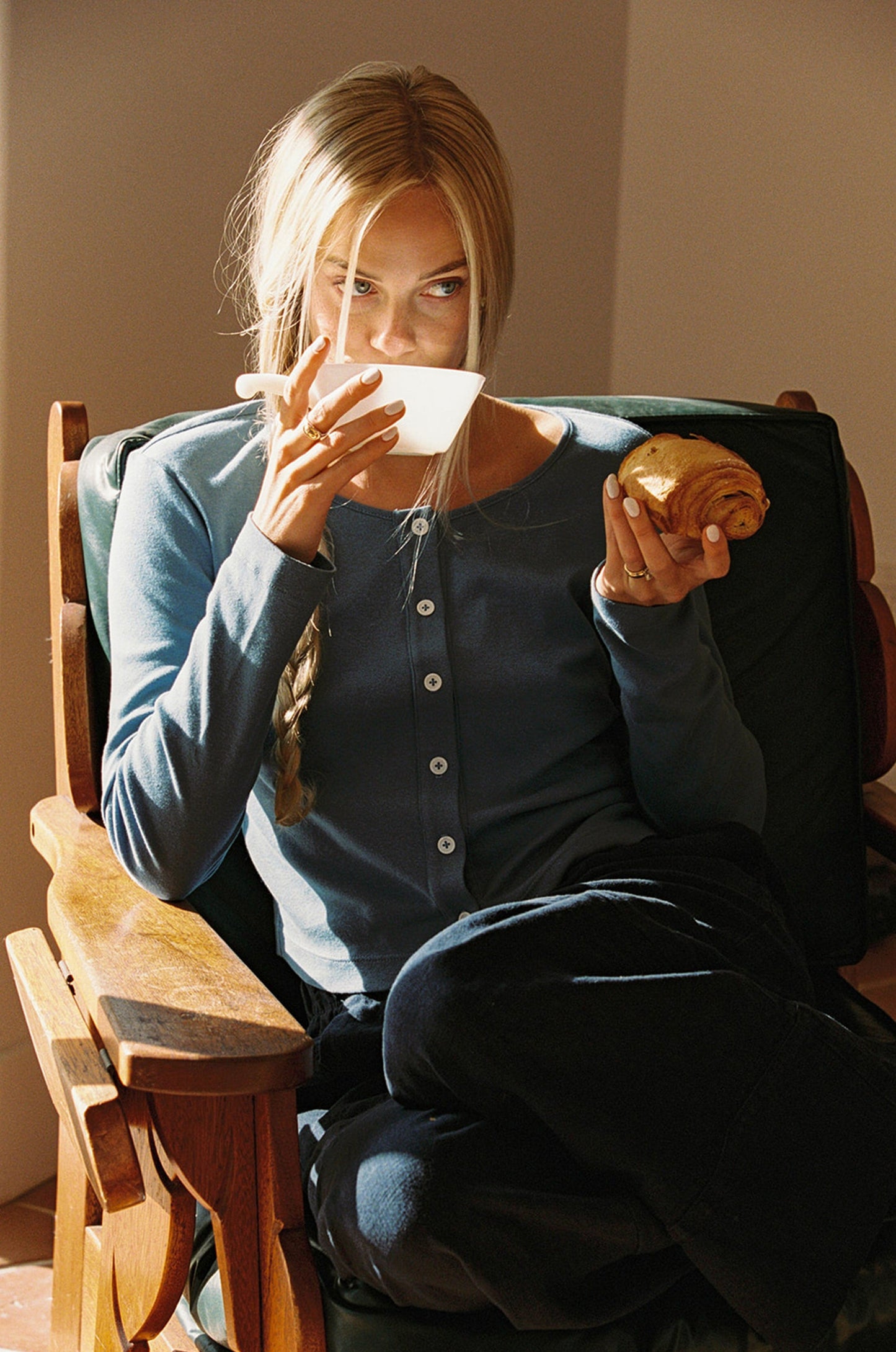 Blue 
A person with long blonde hair sits in a wooden chair, enjoying a croissant and cup. They wear an Organic Rib Cardigan by LA Relaxed and dark pants, as sunlight casts warm shadows across the scene.