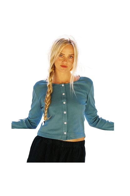 A person with long blonde hair in a braid wears the LA Relaxed Organic Rib Cardigan, featuring pearlescent buttons, paired with dark pants. They stand with arms slightly out to the sides against a completely blank background.