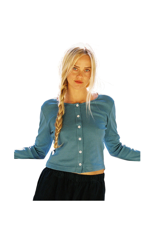 A person with long blonde hair in a braid wears the LA Relaxed Organic Rib Cardigan, featuring pearlescent buttons, paired with dark pants. They stand with arms slightly out to the sides against a completely blank background.