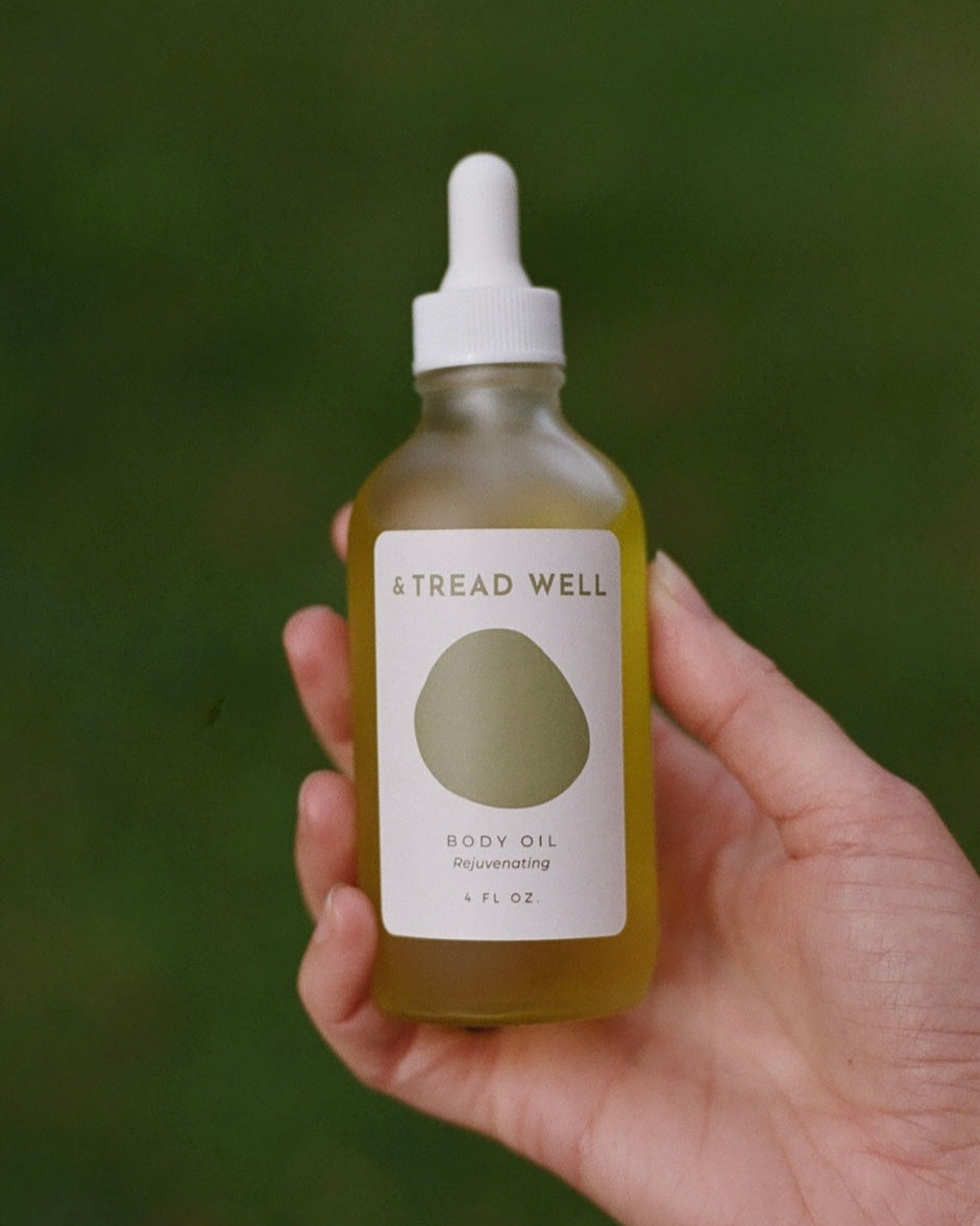 A hand holds a & Tread Well Body Oil glass dropper bottle against a blurred green background, ideal for hydrating your skin and supporting its natural barrier.