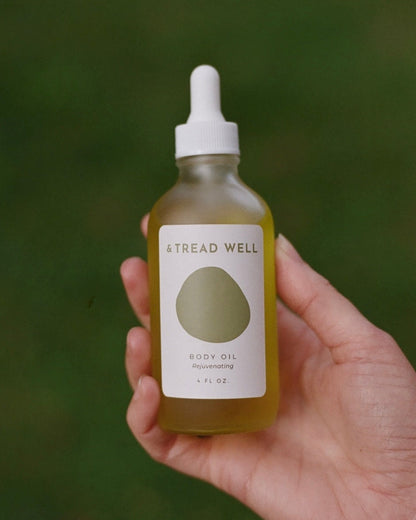 A hand holds a & Tread Well Body Oil glass dropper bottle against a blurred green background, ideal for hydrating your skin and supporting its natural barrier.