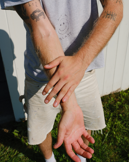 A person outdoors presses one hand to their tattooed forearm, wearing a light gray t-shirt and shorts on grassy ground—ideal skin for hydrating with & Tread Well Body Oil to lock in moisture and support a natural barrier.