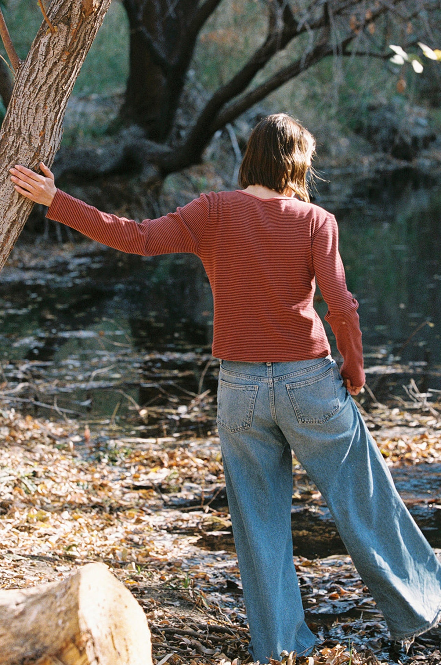 Wearing an LA RELAXED Organic Stripe Long Sleeve and blue jeans, a person leans against a tree by a forest pond. The ground is carpeted with dry leaves, surrounded by trees and still water. Facing away from the camera, they blend seamlessly into the tranquil scene.