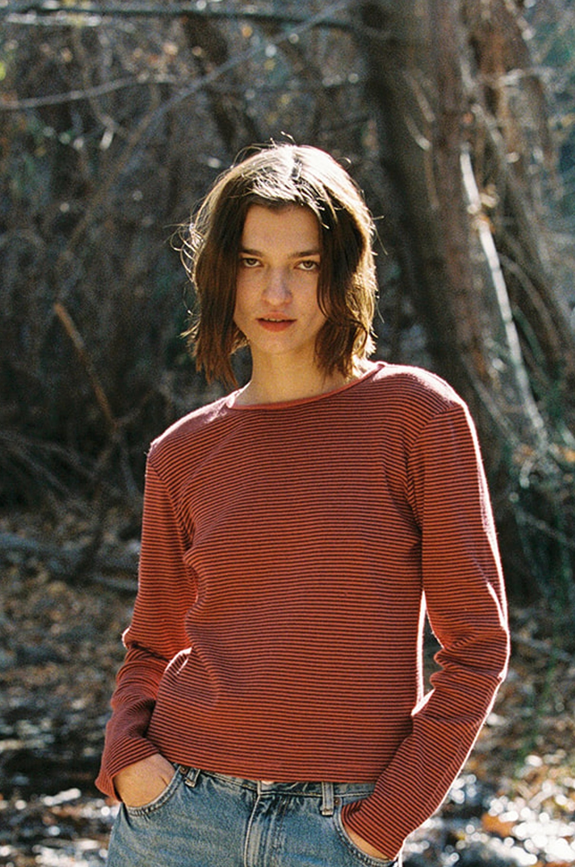 In a sunlit wooded area, a person with short brown hair wears LA RELAXED's Organic Stripe Long Sleeve shirt in red and black, paired with jeans. Their hands rest in their pockets as they gaze at the camera.