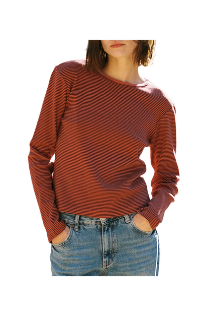 In front of a plain background, a person wears LA RELAXED's Organic Stripe Long Sleeve, featuring red and black stripes crafted from organic cotton, paired with blue jeans. Hands rest casually in pockets; face not visible.