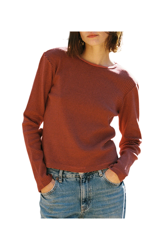 In front of a plain background, a person wears LA RELAXED's Organic Stripe Long Sleeve, featuring red and black stripes crafted from organic cotton, paired with blue jeans. Hands rest casually in pockets; face not visible.