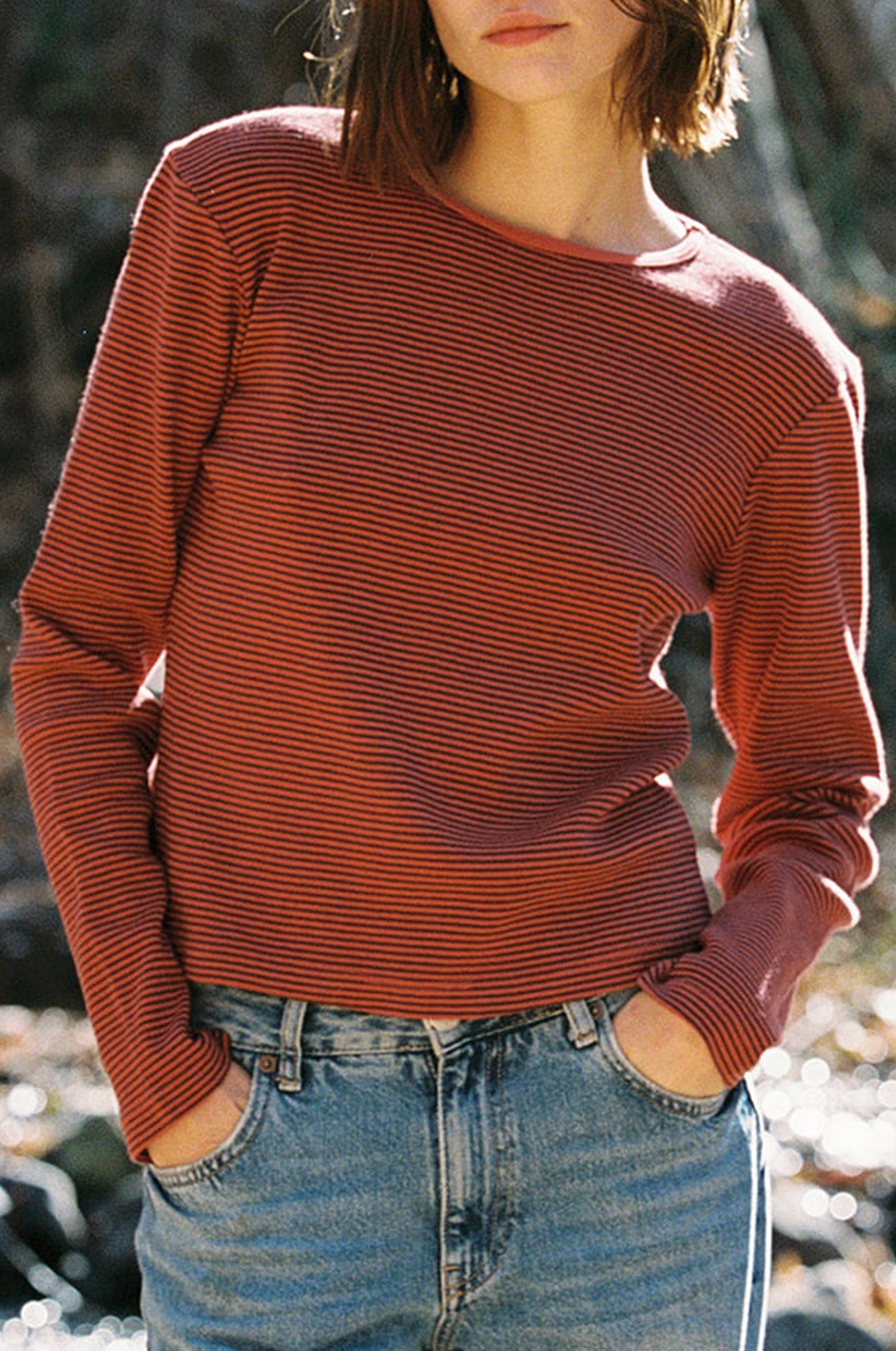 Someone stands with hands in pockets, wearing the LA RELAXED Organic Stripe Long Sleeve, a red and black striped cropped tee made of organic cotton, paired with light blue jeans. Trees and natural scenery create a blurred background.