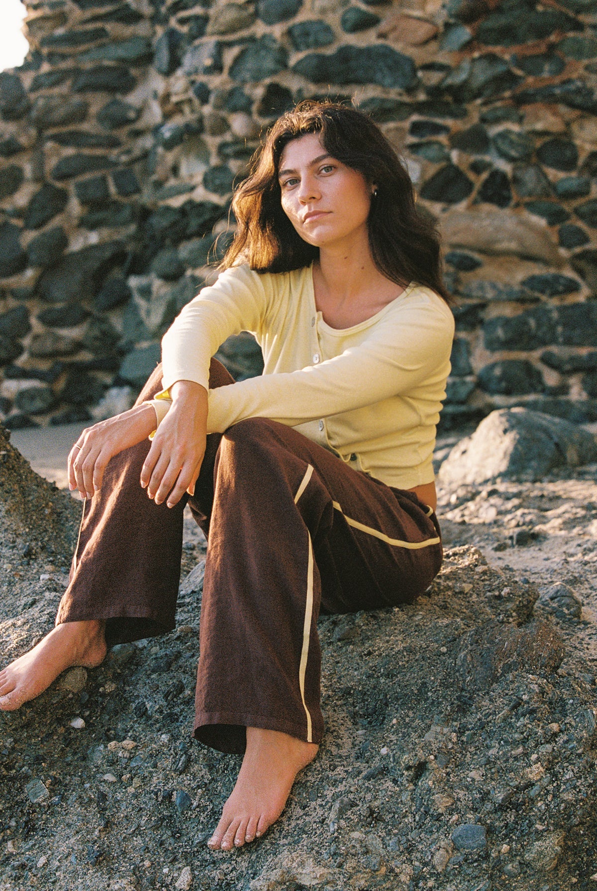 A woman with long dark hair sits barefoot on rocky ground outdoors against a stone wall, wearing LA RELAXED's Linen Track Pant featuring a racing stripe and drawstring waistband, paired with a light yellow cardigan.