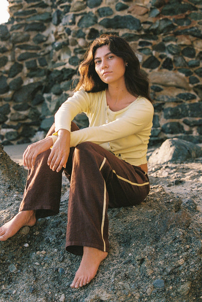 A woman with long dark hair sits barefoot on rocky ground outdoors against a stone wall, wearing LA RELAXED's Linen Track Pant featuring a racing stripe and drawstring waistband, paired with a light yellow cardigan.