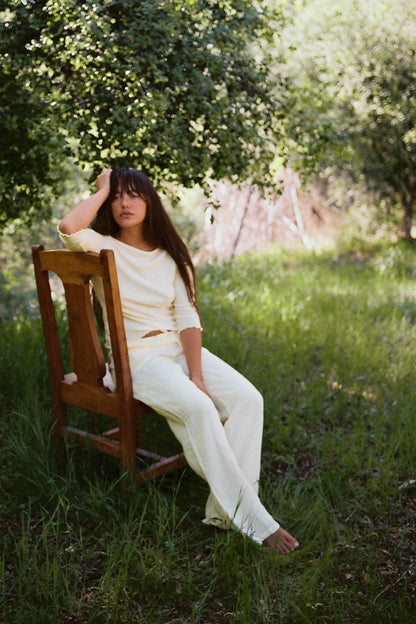 A young person with long dark hair wears LA RELAXED’s Linen Track Pant, cream-colored with a drawstring waist, as they sit barefoot on a wooden chair in a grassy, sunlit outdoor area surrounded by green trees.