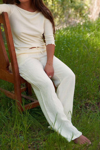 Wearing the LA RELAXED Linen Track Pant with a drawstring waist and a light cream long-sleeve top, someone sits barefoot outdoors on a wooden chair, surrounded by green grass and trees. Their face is not shown.