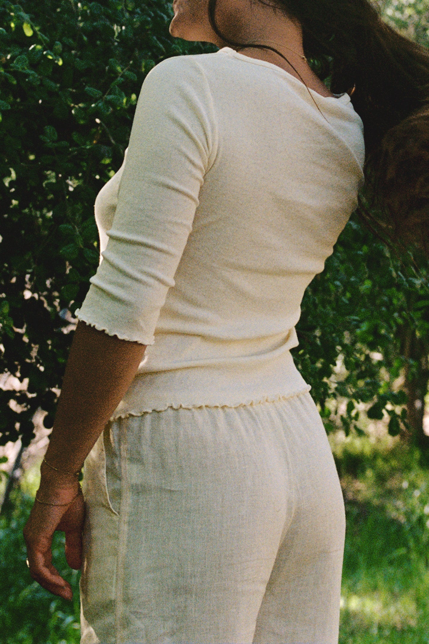 A person stands outdoors near green foliage, facing slightly away from the camera, wearing LA RELAXED Linen Track Pant and a cream long-sleeve top. Their hair is pulled back as sunlight filters through the trees.