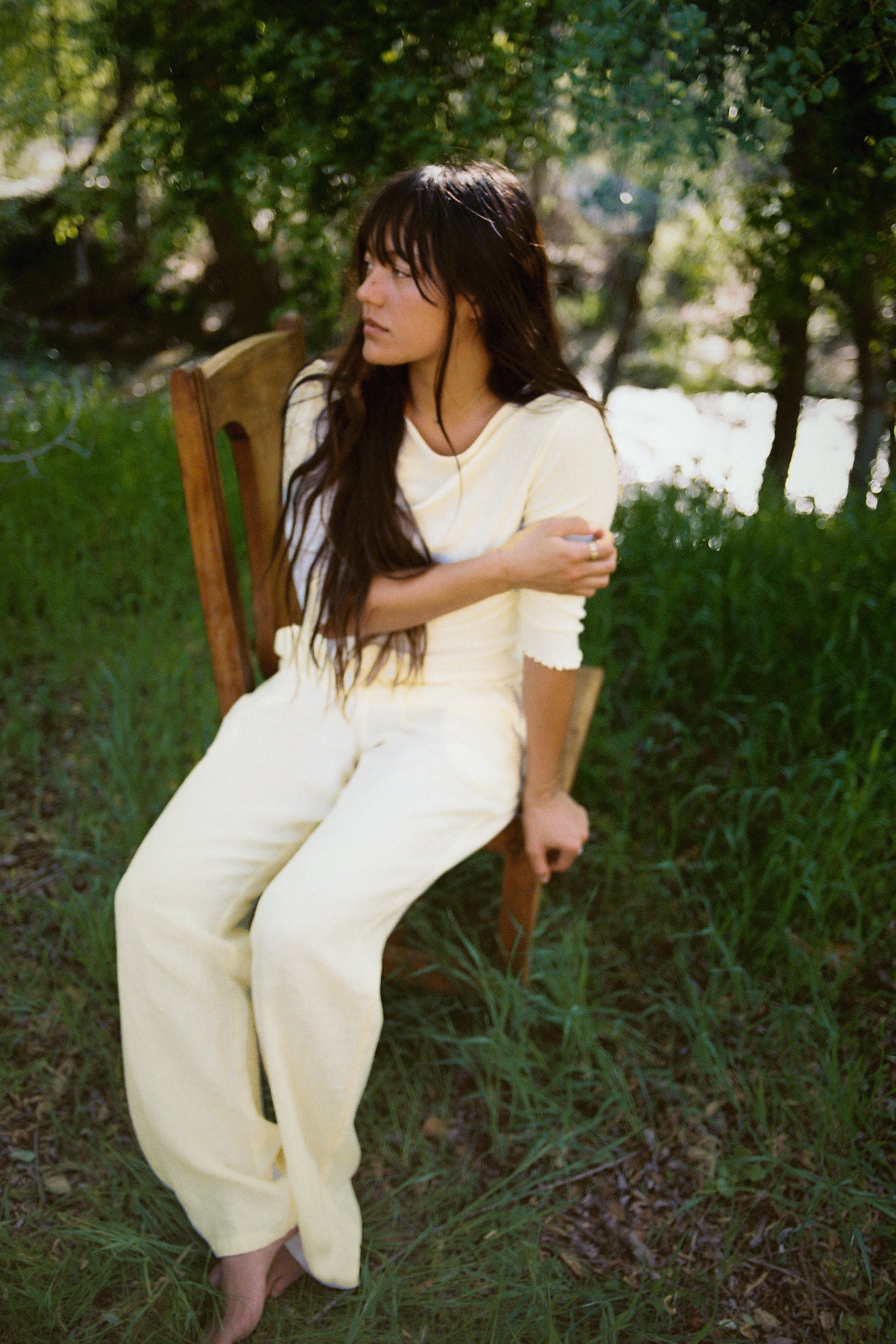 A woman sits barefoot on a wooden chair outdoors in the grass, wearing LA RELAXED Linen Track Pant in light yellow with a drawstring waist. Sunlight streams through the trees as she looks to the side.