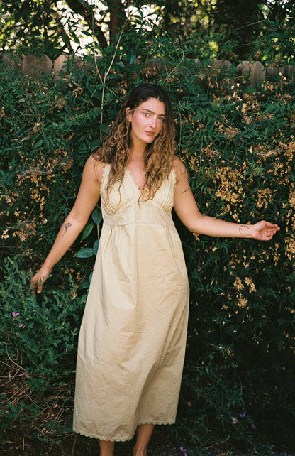Butter - A woman with wavy brown hair and tattoos on both arms stands outdoors before green foliage, wearing the LA RELAXED Makinna Poplin Dress, a light-colored organic poplin style with cotton lace trim and a calm expression.